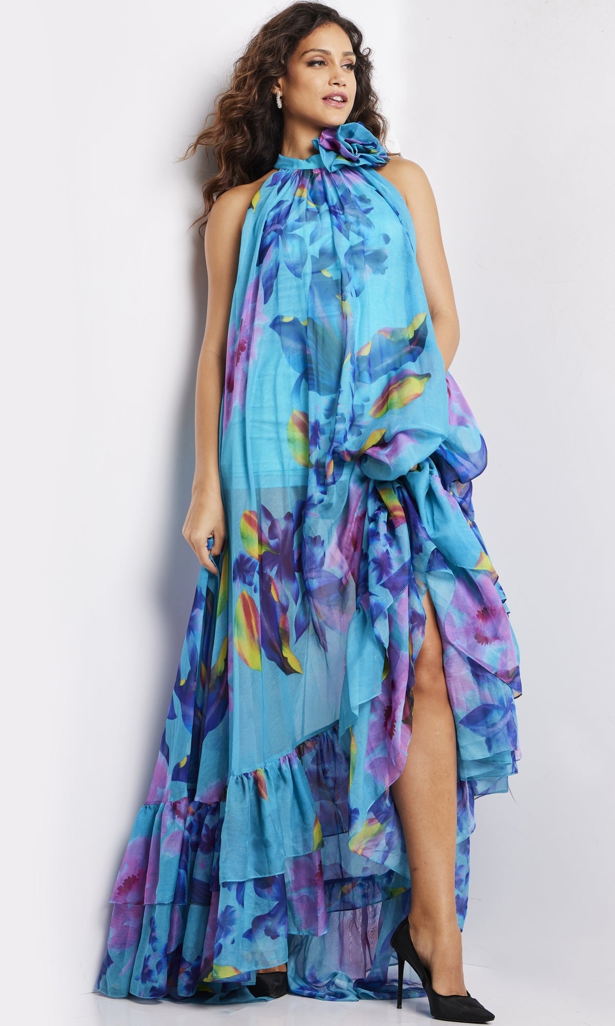 A woman stands against a white background in the Long Prom Dress 38721 by Jovani, featuring a flowing sleeveless blue design with ruffles, a high neckline, high slit, and paired with black heels.